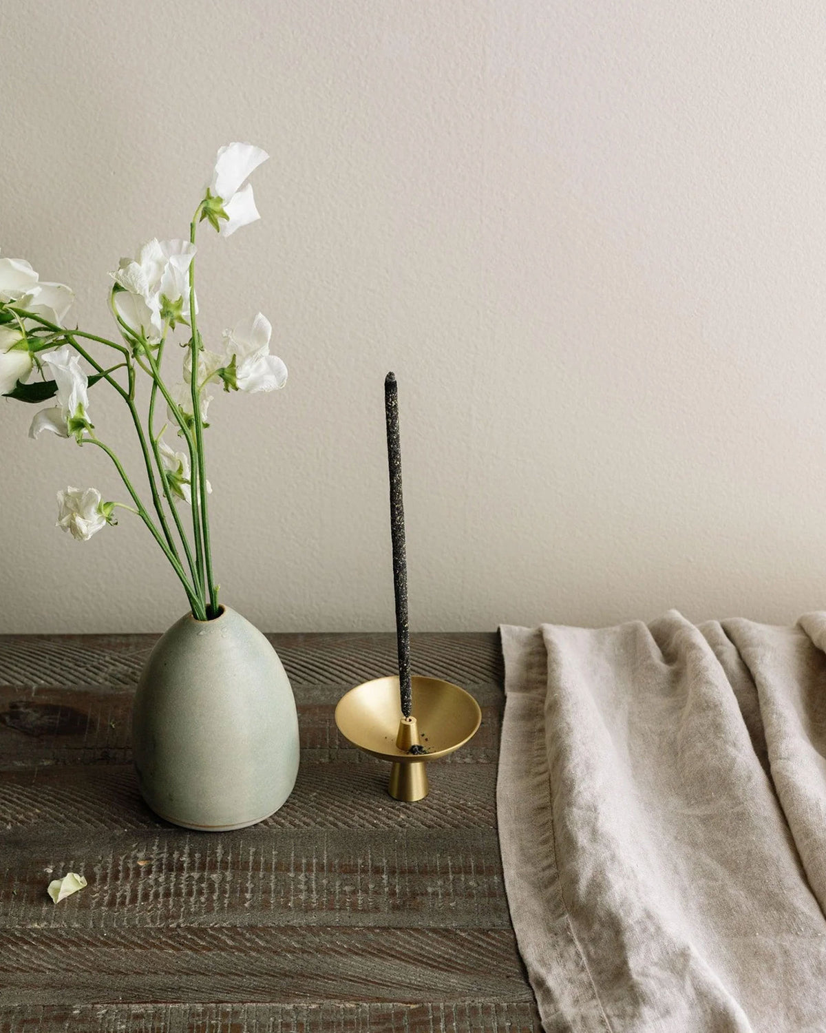 Small brass satellite shaped incense holder with a tall black incense stick standing straight up from the centre. Set on a rustic style wood table next to a white vase with tall white flowers and a piece of light grey linen cloth.