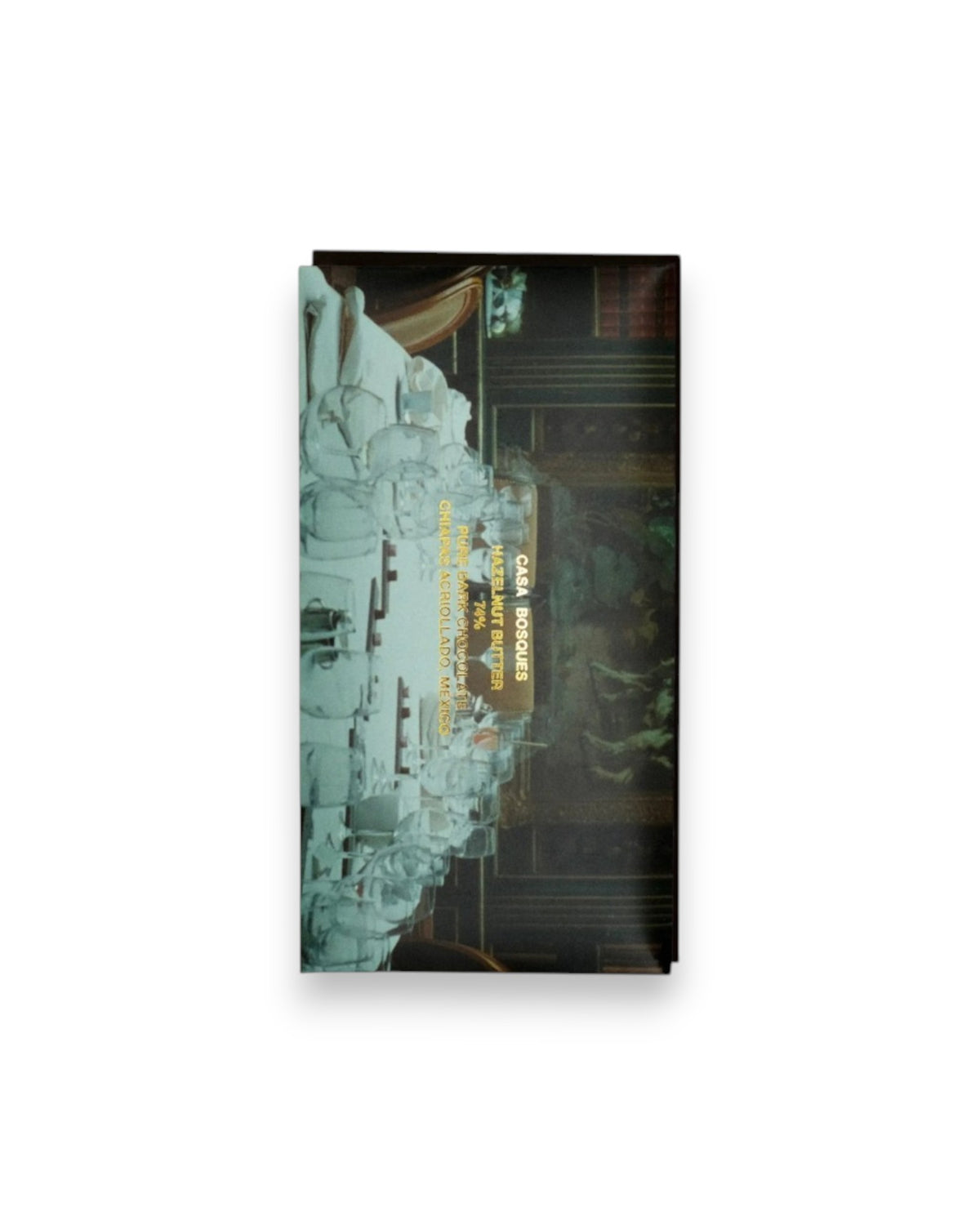 A rectangular packaged bar of Casa Bosques chocolate against a solid white background, label reads "CASA BOSQUES HAZELNUT BUTTER 74%" The graphic on packaging is restaurant table full of clear, unused glassware set against an old roman painting.
