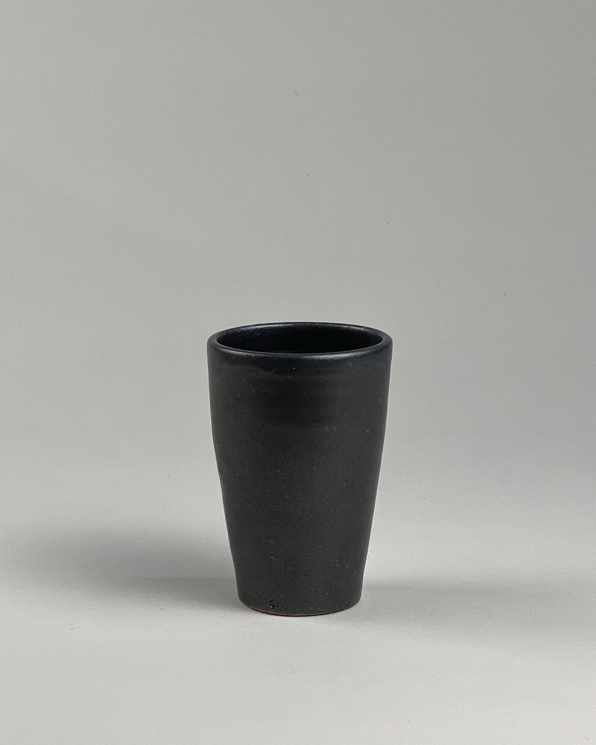 A tall black ceramic drinking tumbler on a seamless, light grey background.