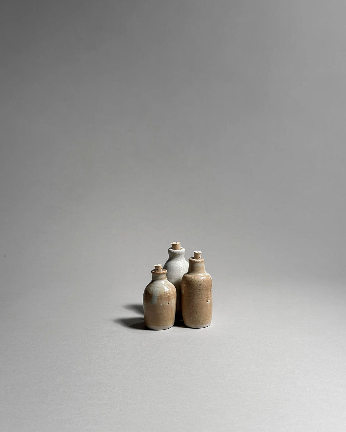 A grouped set of three ceramic bottles with cork lids and a rustic white and brown glaze on a seamless, light grey background.