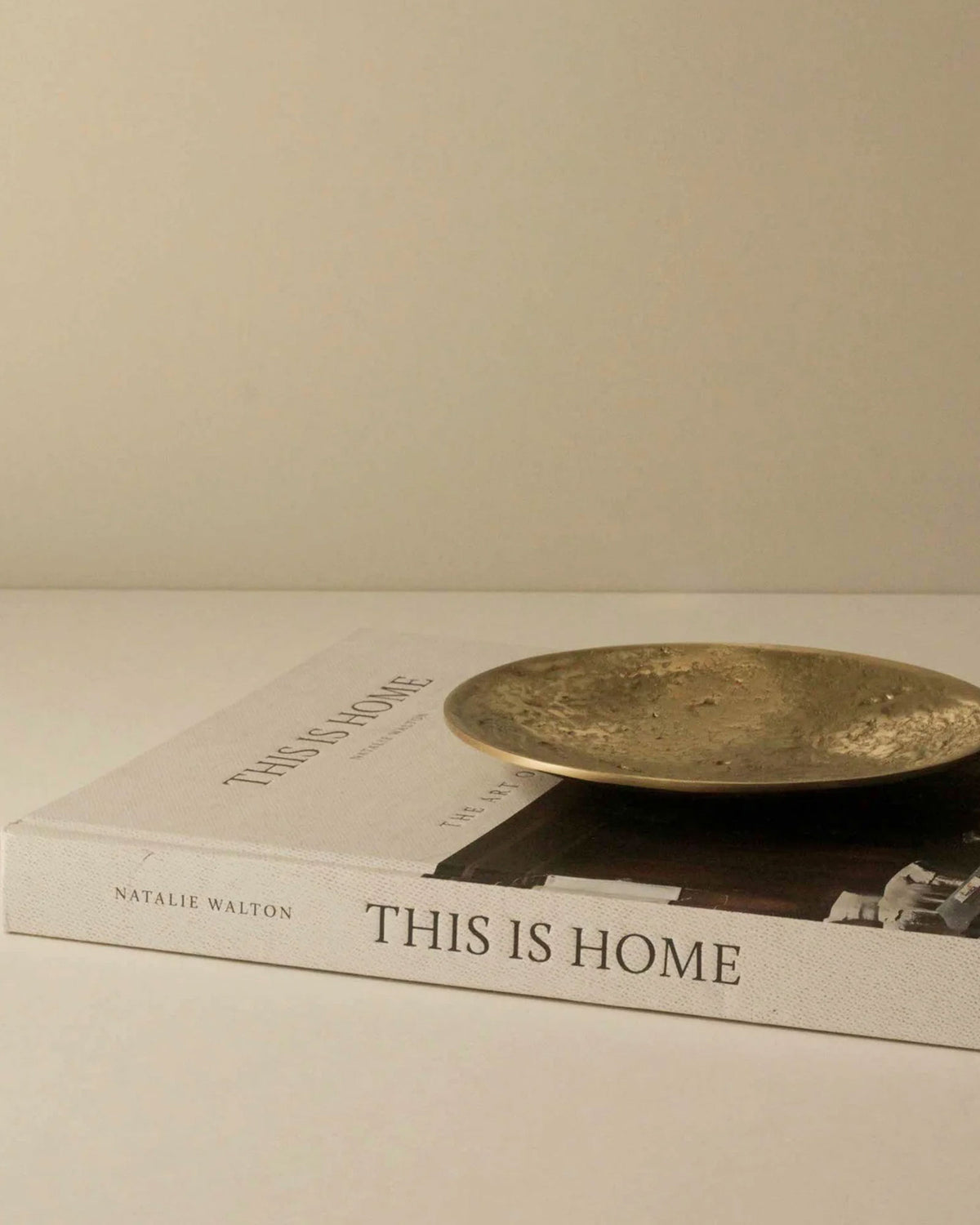 A round brass textured plate sits on top of a large coffee-table book titled This is Home. White table surface and off-white background.