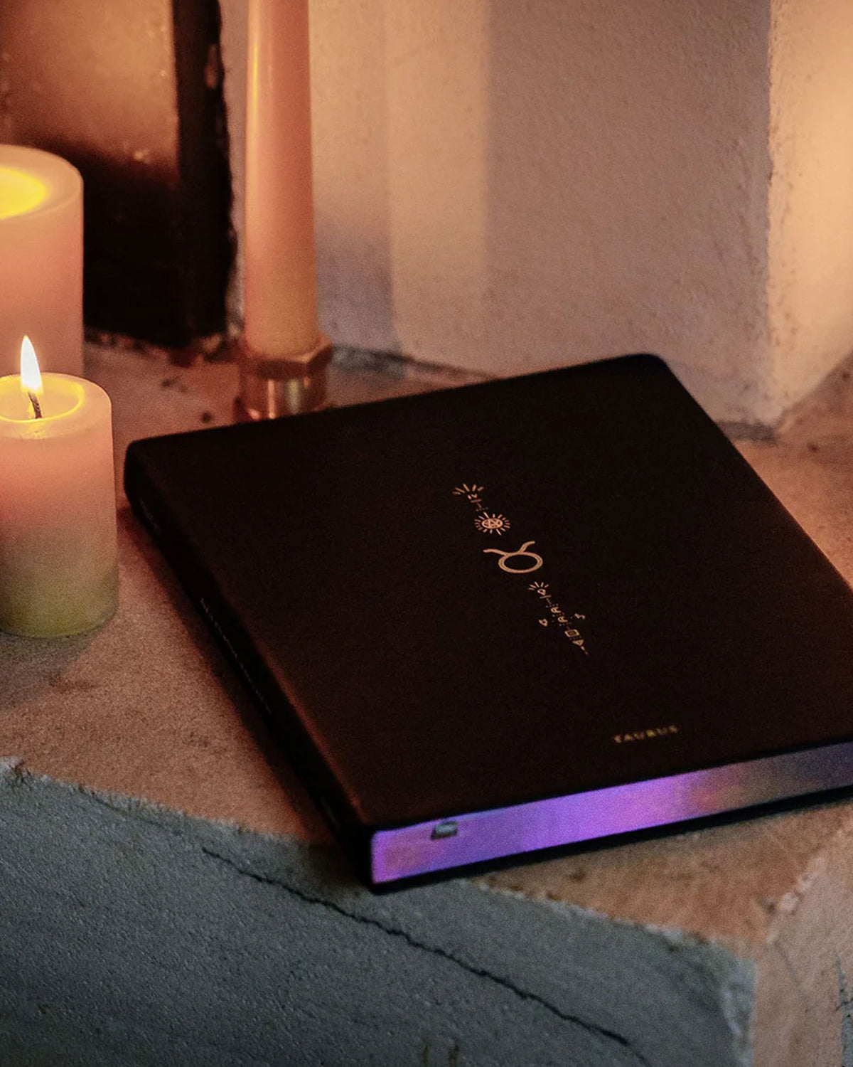 A black hardcover book with zodiac symbols on the cover, styled on a stone ledge next to lit candles in a warm, atmospheric setting.