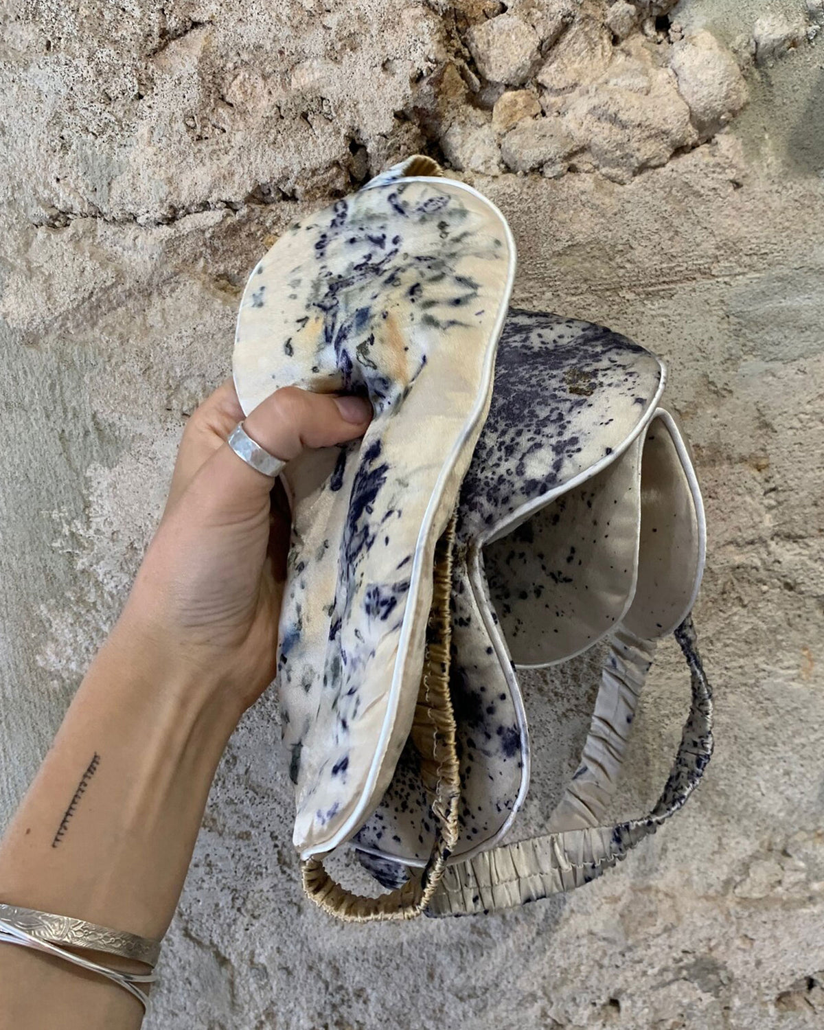 A hand holds a bunch of three silk eye masks with a cream base and dark blue speckled patterns, set against a textured stone wall.