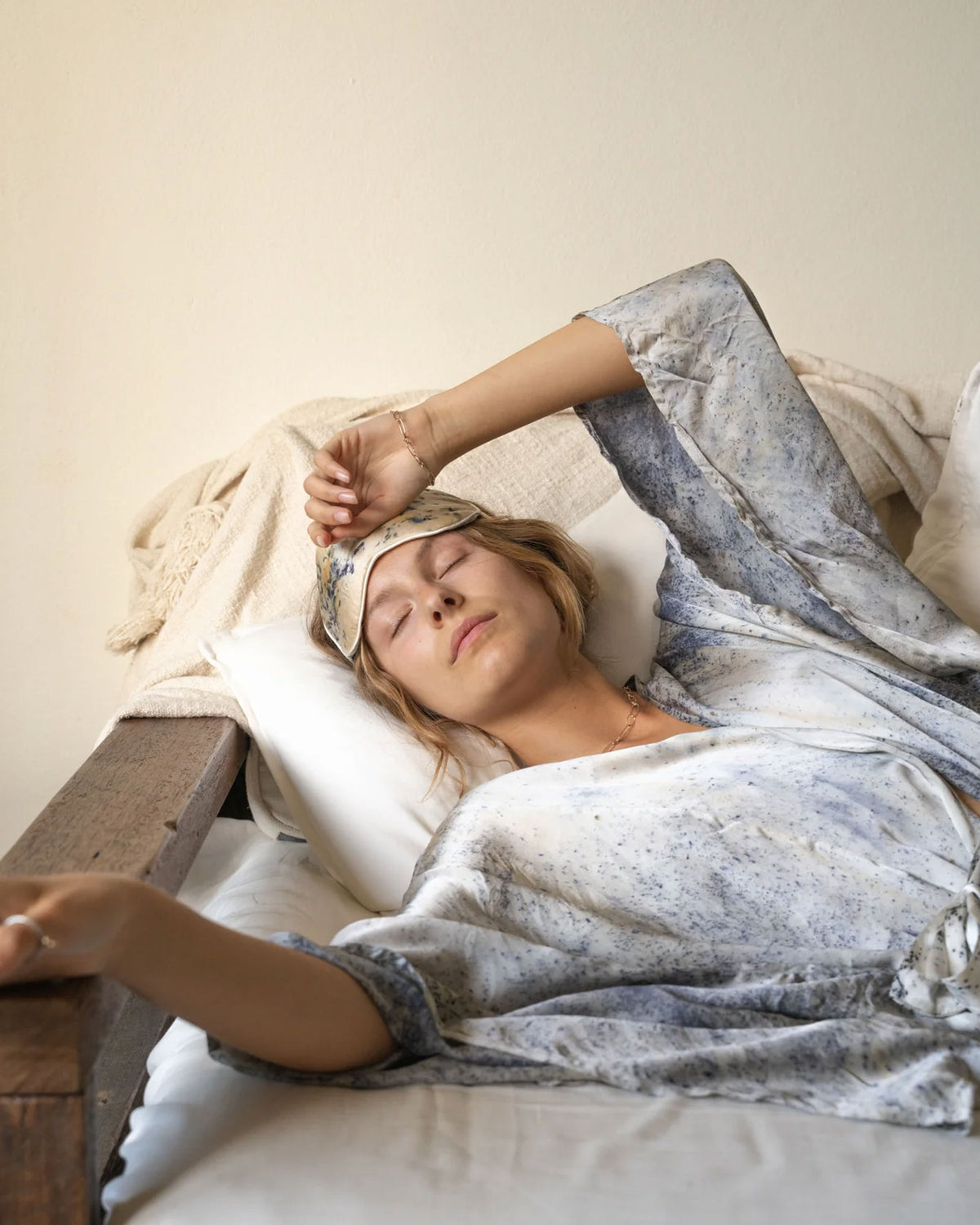 A woman lying on a bed with her eyes closed, wearing a blue-gray patterned robe and a matching eye mask that is lifted above her eyes. The room has a warm cream-colored wall and white bedding.