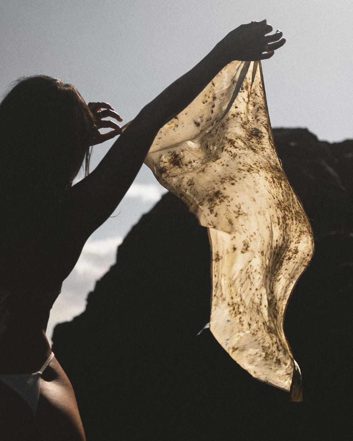 Silhouette of a woman holding up a brown mottled scarf in the wind. Set against an outdoor, rocky backdrop.
