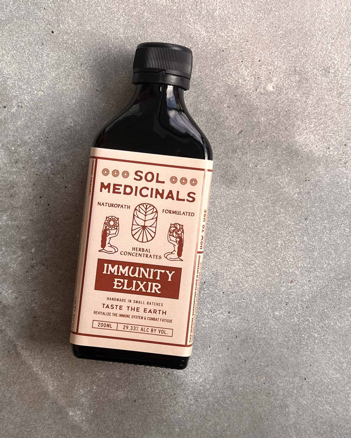 A bottle of Sol Medicinals Immunity Elixir on a textured concrete surface. The product is in an amber glass bottle with a black cap and a cream-coloured label with red and cream text.