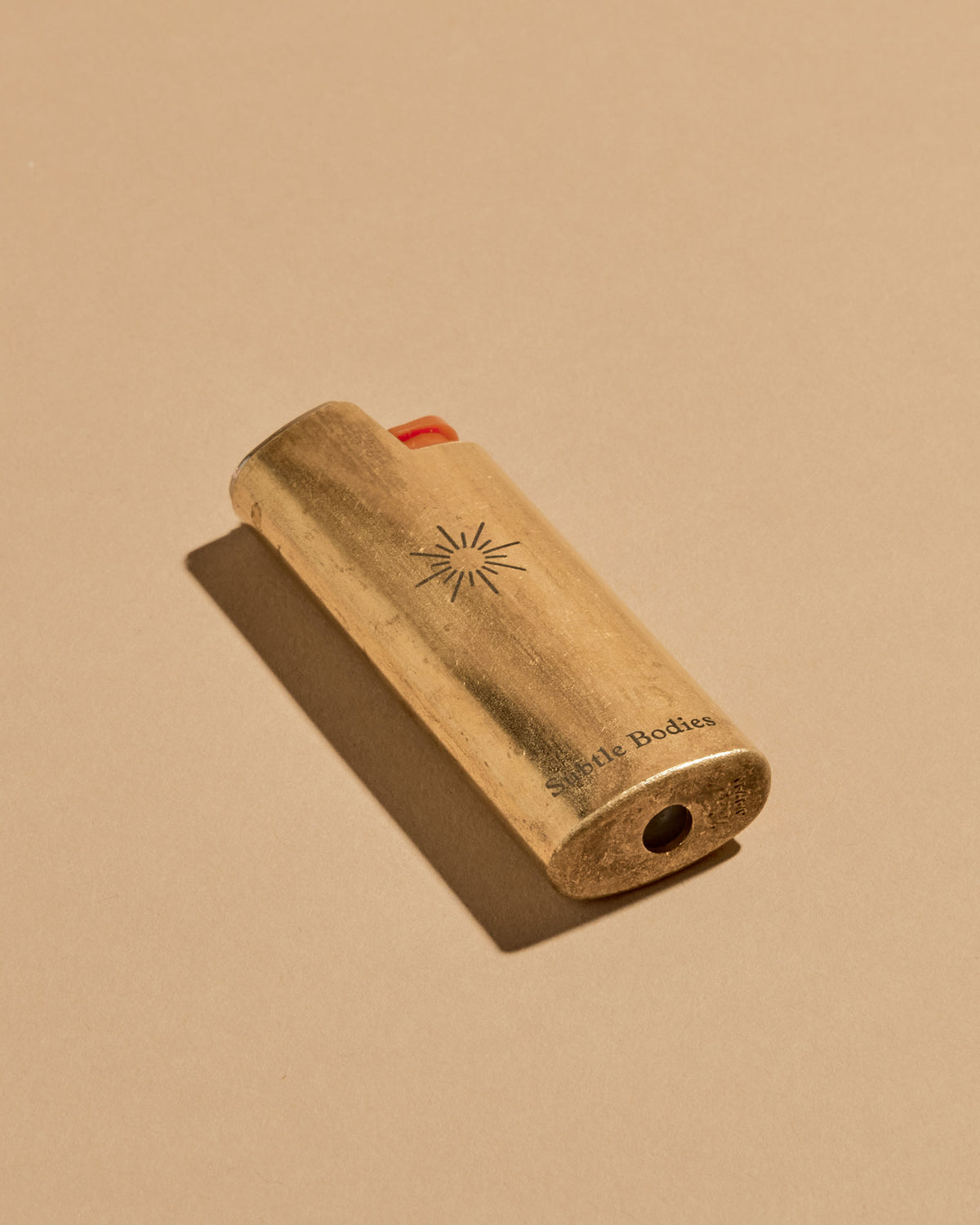 A mini BIC lighter in a brass lighter case with a matte finish. A geometric sun and the Subtle Bodies logo is etched on to the front of the lighter case. Set on a seamless, light orange background.