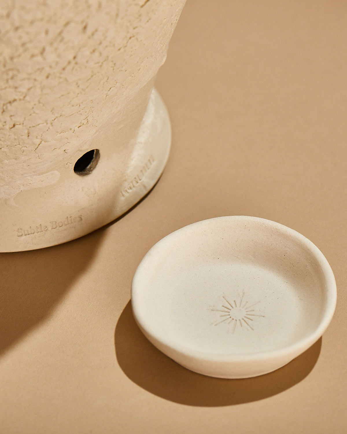 A small white ceramic unglazed dish with the impression of a sun is on a light orange surface next to another ceramic object with a textured surface and small circular hole at the base.