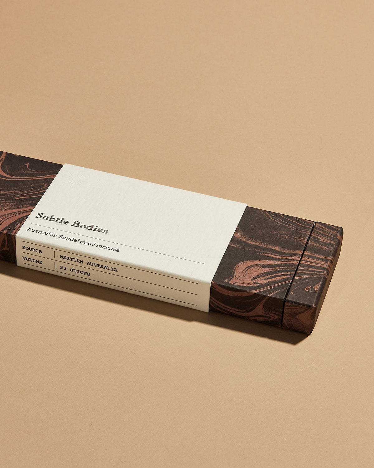 A closed red marbled box of Subtle Bodies Australian Sandalwood Incense with a white wrap-around label. Set on a light orange background.