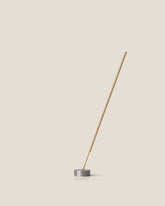 A light brown incense stick stands vertically, tilting to the right, from the centre of a small round, silver incense holder on an off-white background.