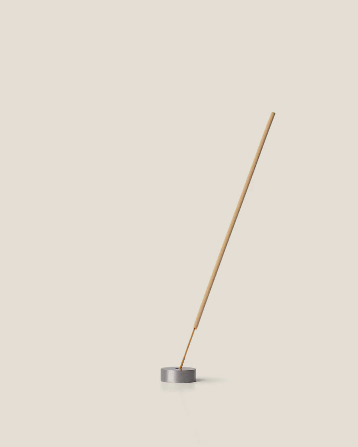 A light brown incense stick stands vertically, tilting to the right, from the centre of a small round, silver incense holder on an off-white background.