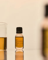 A small clear glass bottle with black screw cap and a translucent brown liquid. Set on a reflective surface with other vessels containing a variation of the coloured liquid. 