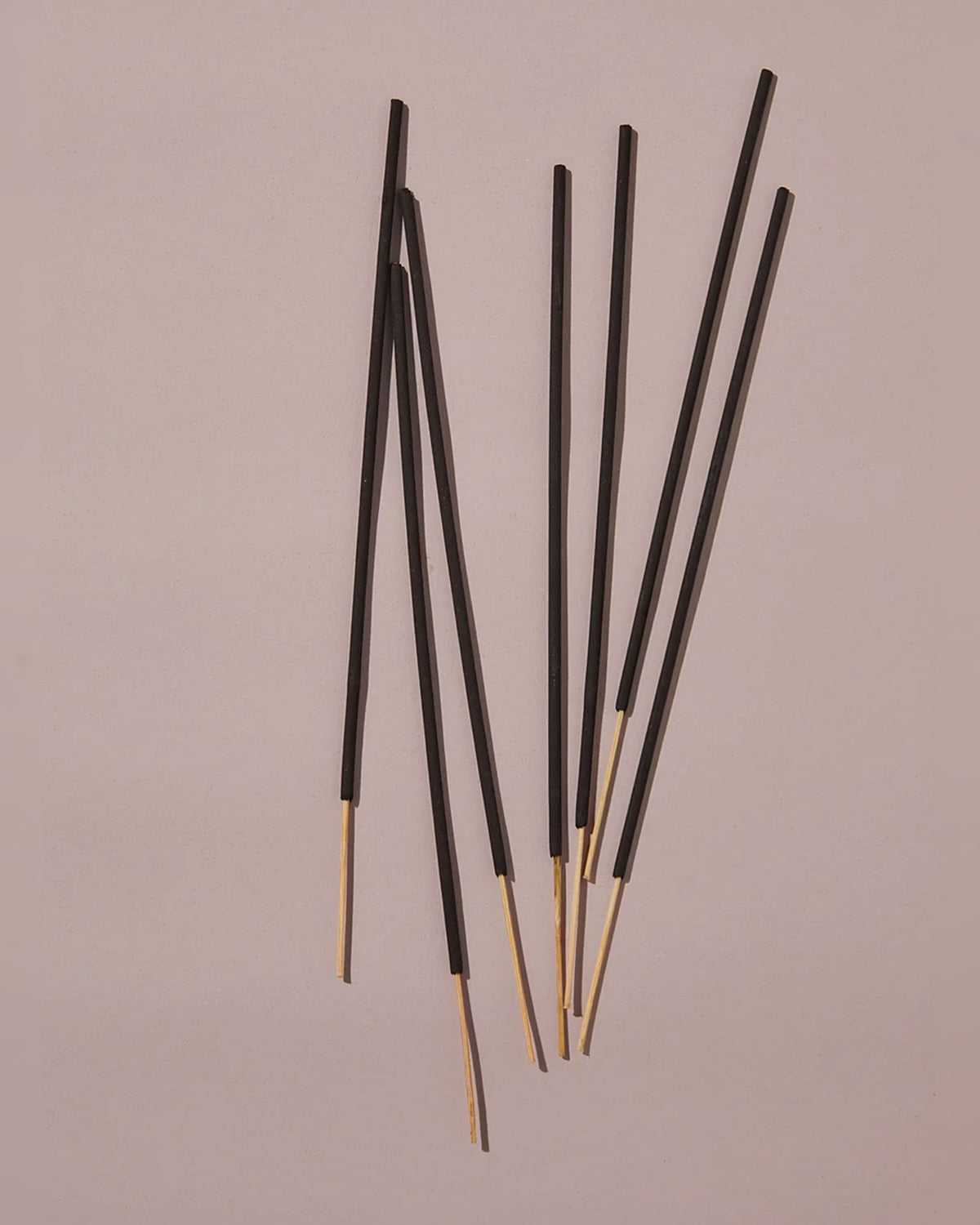 Seven black incense bamboo sticks laying on a dusty-pink background with light and shadow.