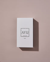A rectangular white AYU NOOR perfume box on a dusty pink background.