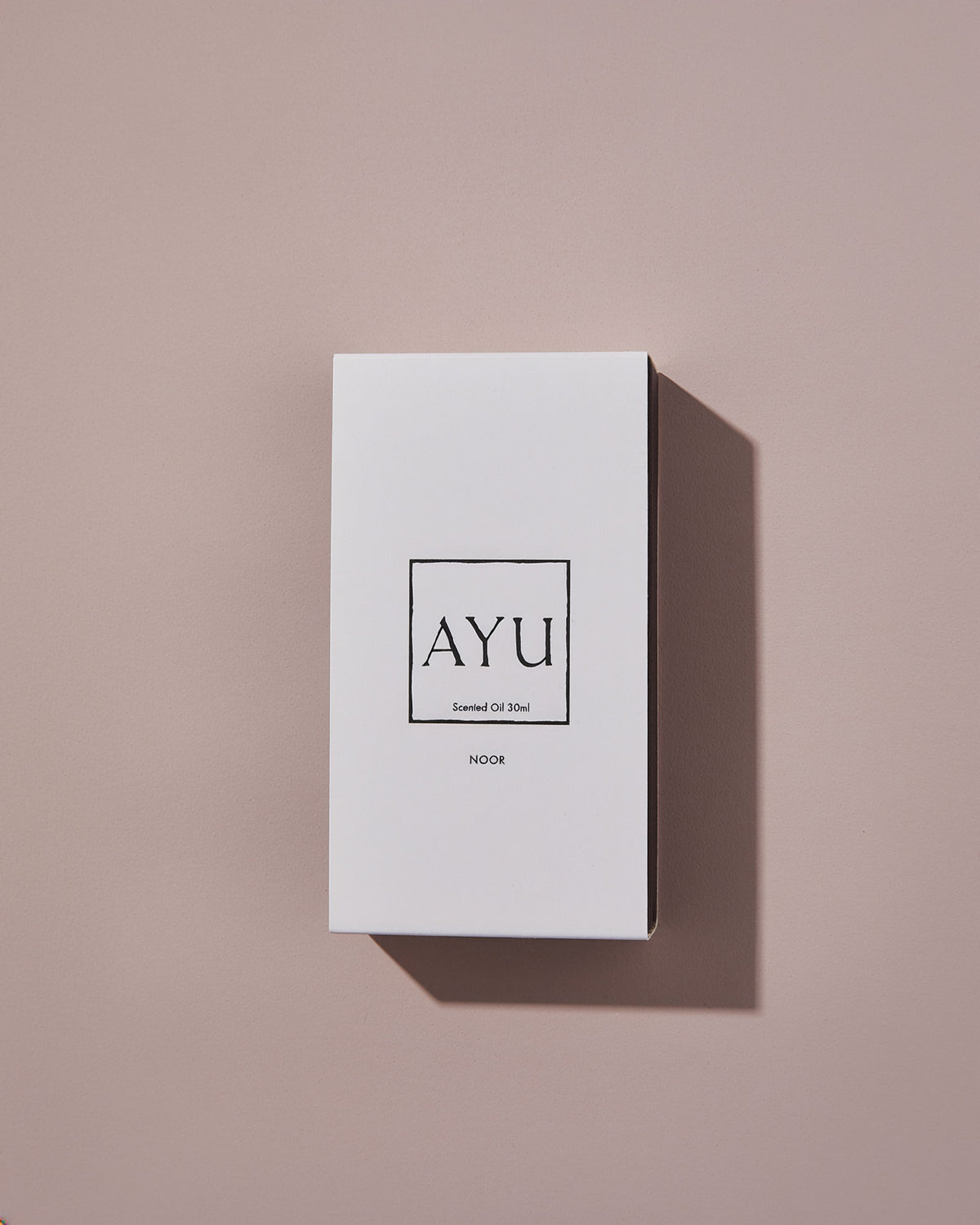 A rectangular white AYU NOOR perfume box on a dusty pink background.