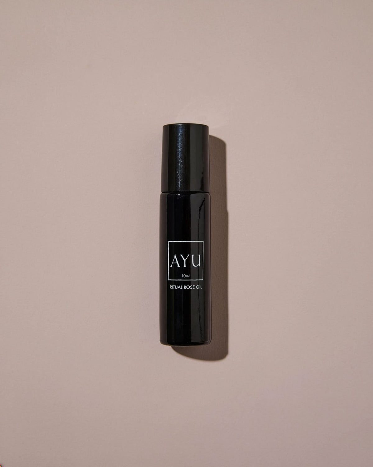 A black cylindrical roll-on bottle of AYU Rose Oil against a dusty pink background.