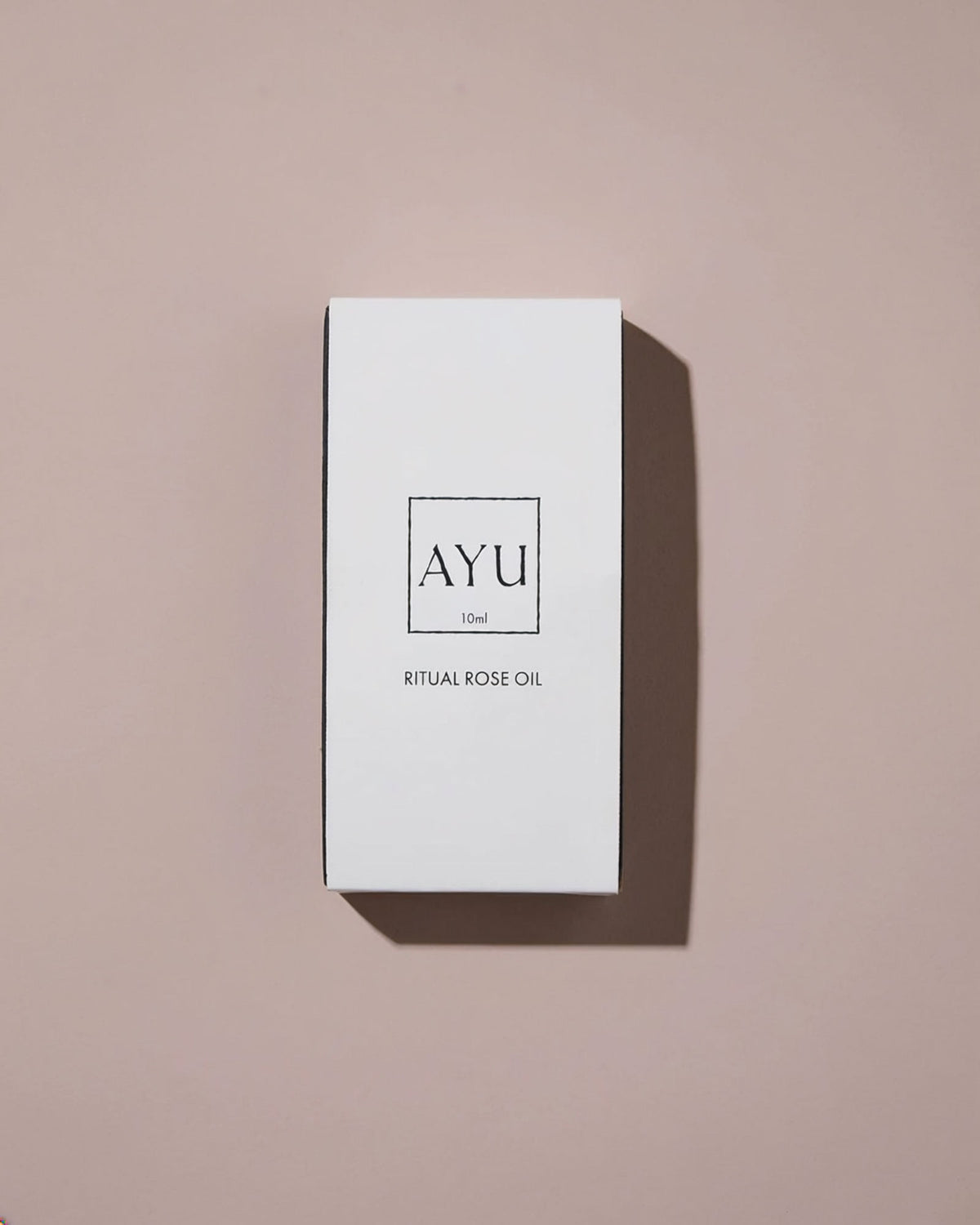 A minimalist white rectangular box for AYU Ritual Rose Oil, featuring a clean black logo and text, set against a solid dusty pink background with a soft shadow.