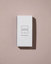 A minimalist white rectangular box for AYU Ritual Rose Oil, featuring a clean black logo and text, set against a solid dusty pink background with a soft shadow.