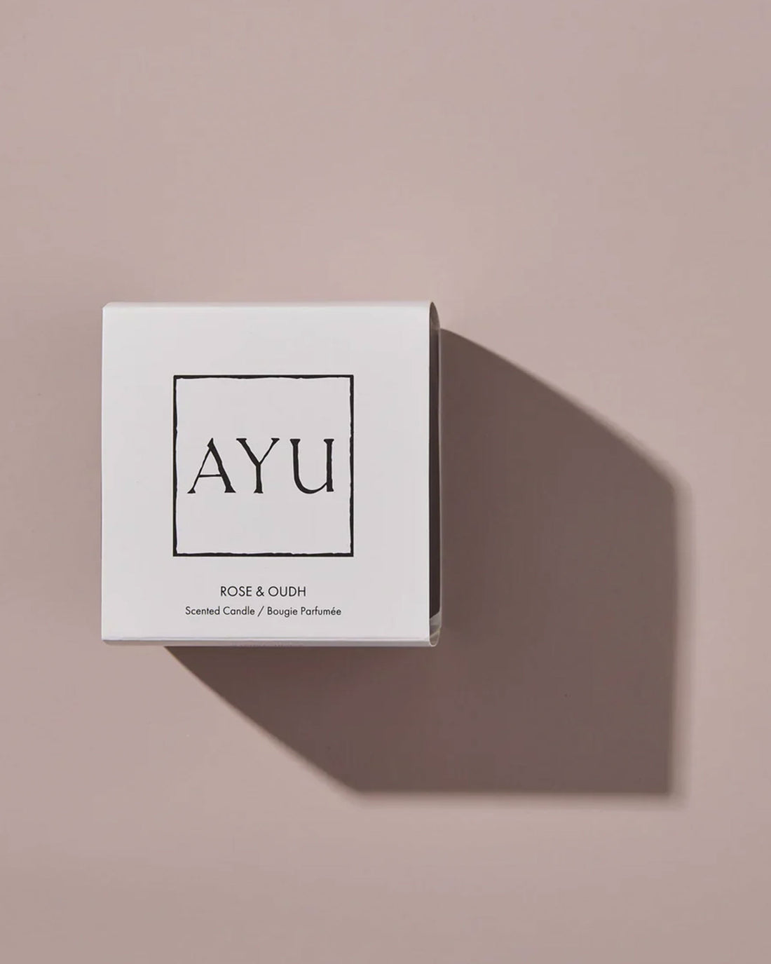 A white cube candle box with black lettering that reads AYU ROSE &amp; OUDH, Scented Candle / Bougie Parfumée on a dusty pink background with dramatic shadows.