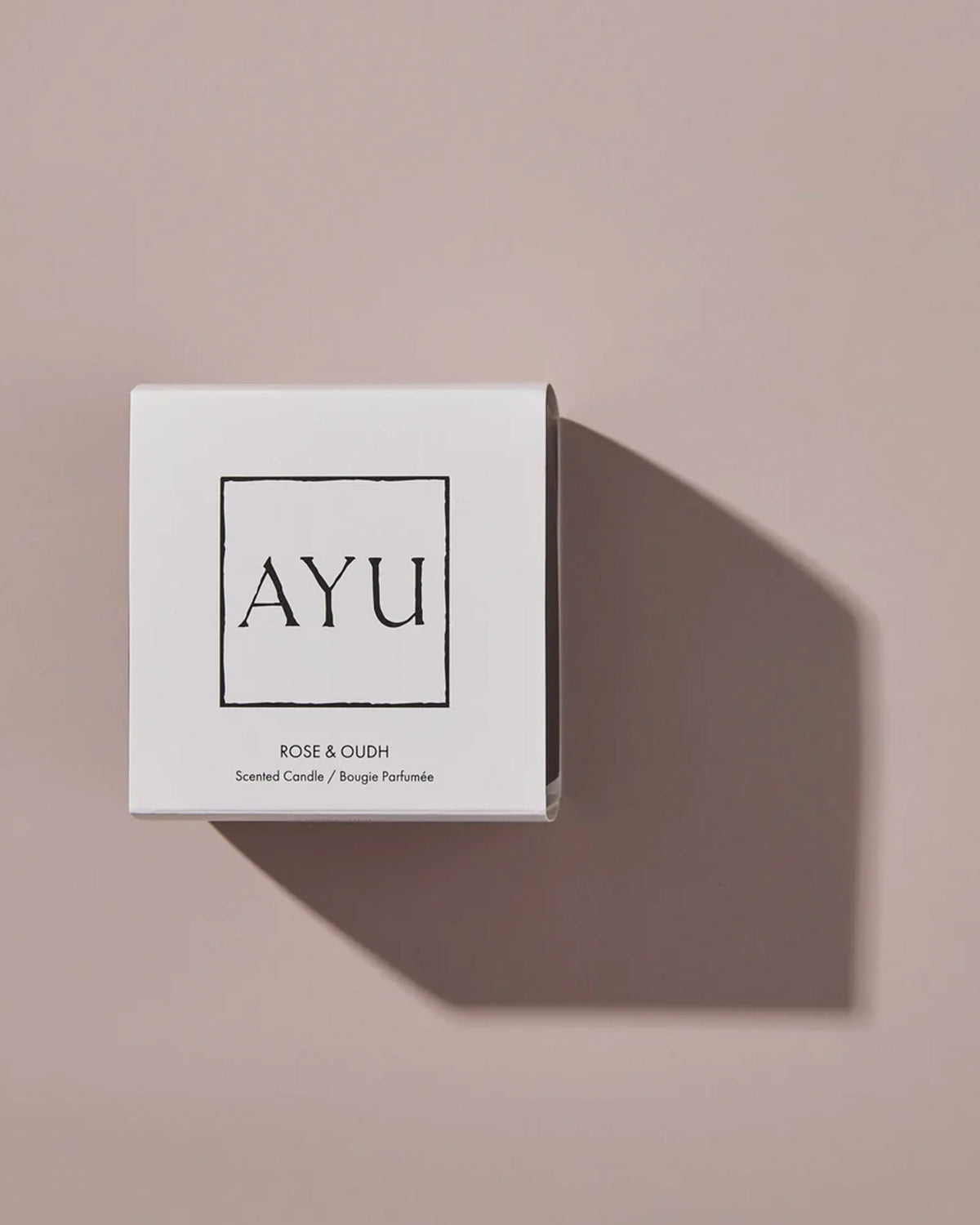 A white cube candle box with black lettering that reads AYU ROSE &amp; OUDH, Scented Candle / Bougie Parfumée on a dusty pink background with dramatic shadows.