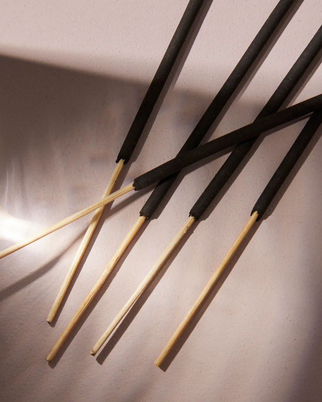 Up-close of five incense sticks laying on a dusty-pink background with light and shadow.