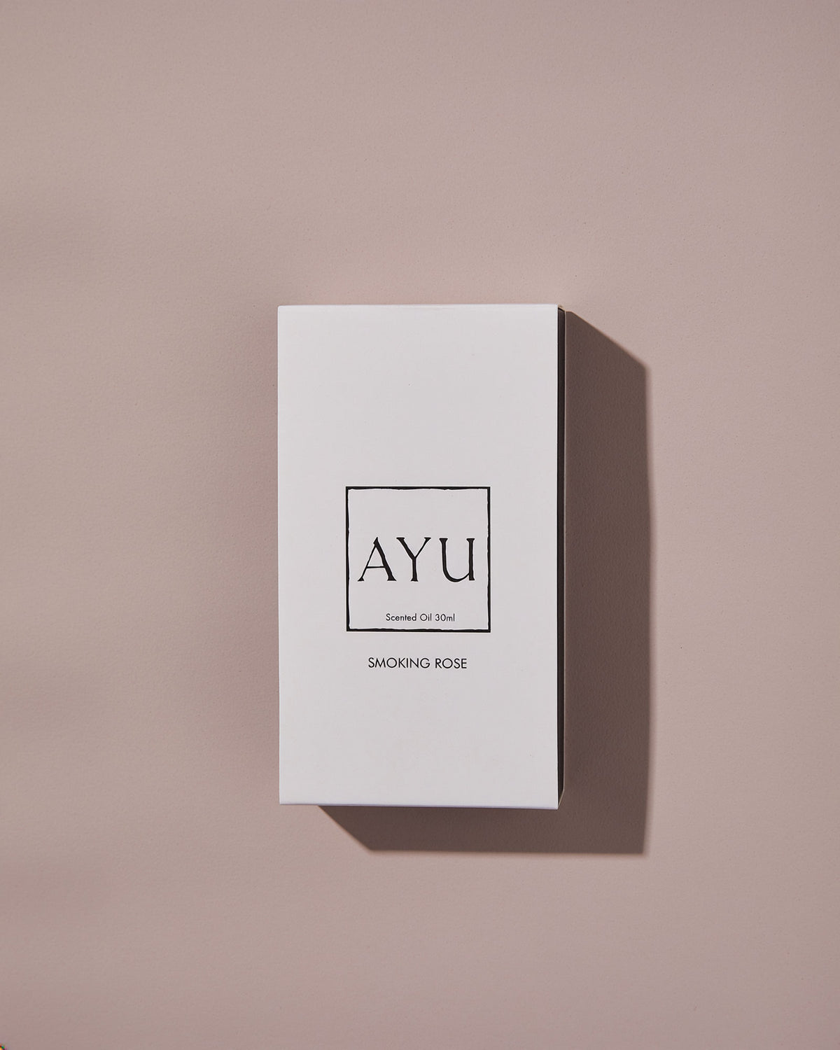 A rectangular white AYU SMOKING ROSE perfume box on a dusty pink background.