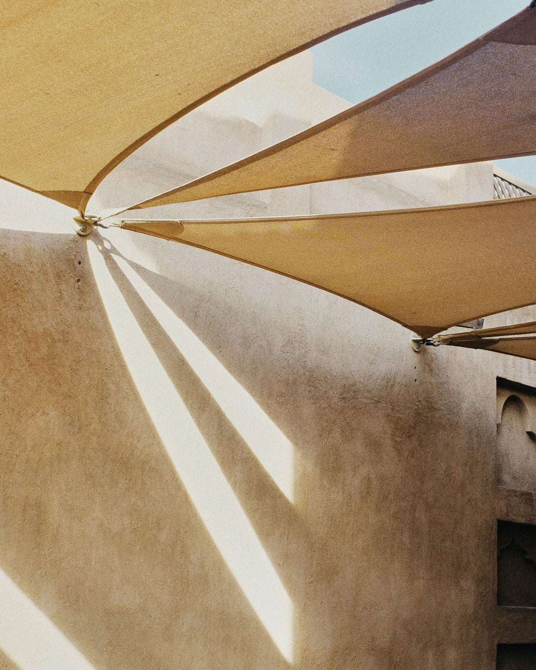 A yellow shade cloth is pinned to a beige textured wall. The sunlight casts shadows of the shapes through the shade cloth. 
