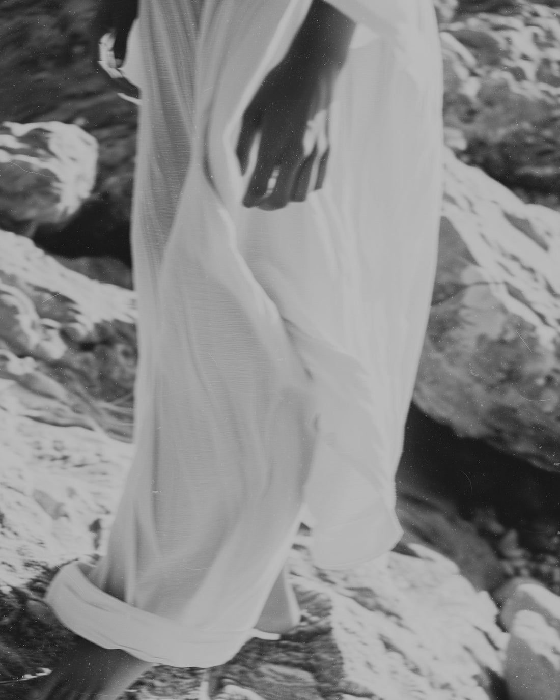 A black and white photograph showing a person from the waist down wearing a loose, flowing white garment while standing on rocky ground.