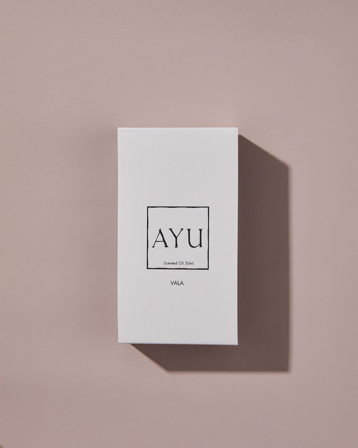 A rectangular white AYU VALA perfume box on a dusty pink background.