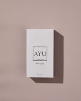 A rectangular white AYU WHITE OUDH perfume box on a dusty pink background.
