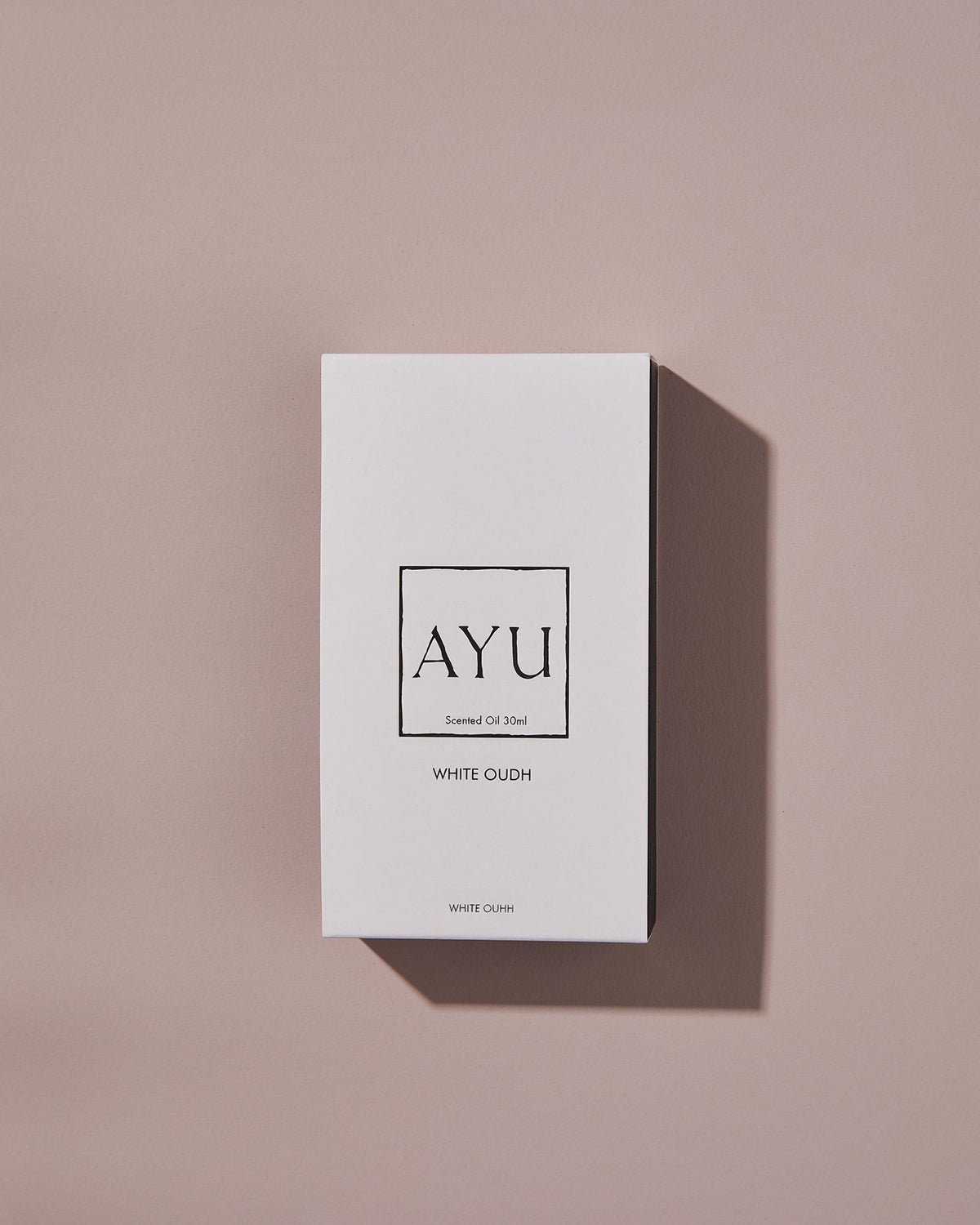 A rectangular white AYU WHITE OUDH perfume box on a dusty pink background.