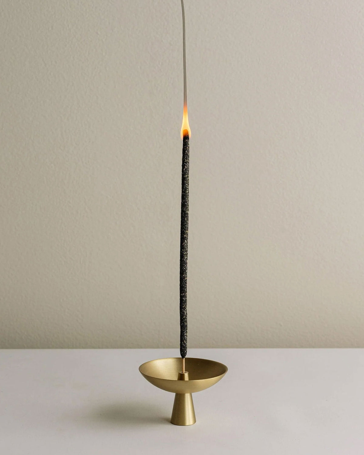 Small brass satellite shaped incense holder with a tall black incense stick standing straight up from the centre. The tip of the incense is lit with a flame and a stream of smoke. Set on a white table surface and off-white background.