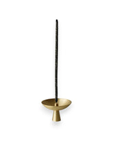 Small brass satellite shaped incense holder with a tall black incense stick standing straight up from the centre. Set against solid white background. 