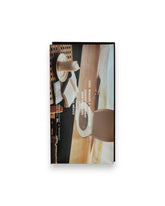 A rectangular packaged bar of Casa Bosques chocolate against a solid white background, label reads "CASA BOSQUES & APARTAMENTO TOASTED BUCKWHEAT DARK CHOCOLATE 74%" The graphic on packaging is a full bleed photo of a retro style toilet recessed into a wooden tabletop bench, the corner of a table is in-shot with a candle on a plate, an open book and a closed book. 