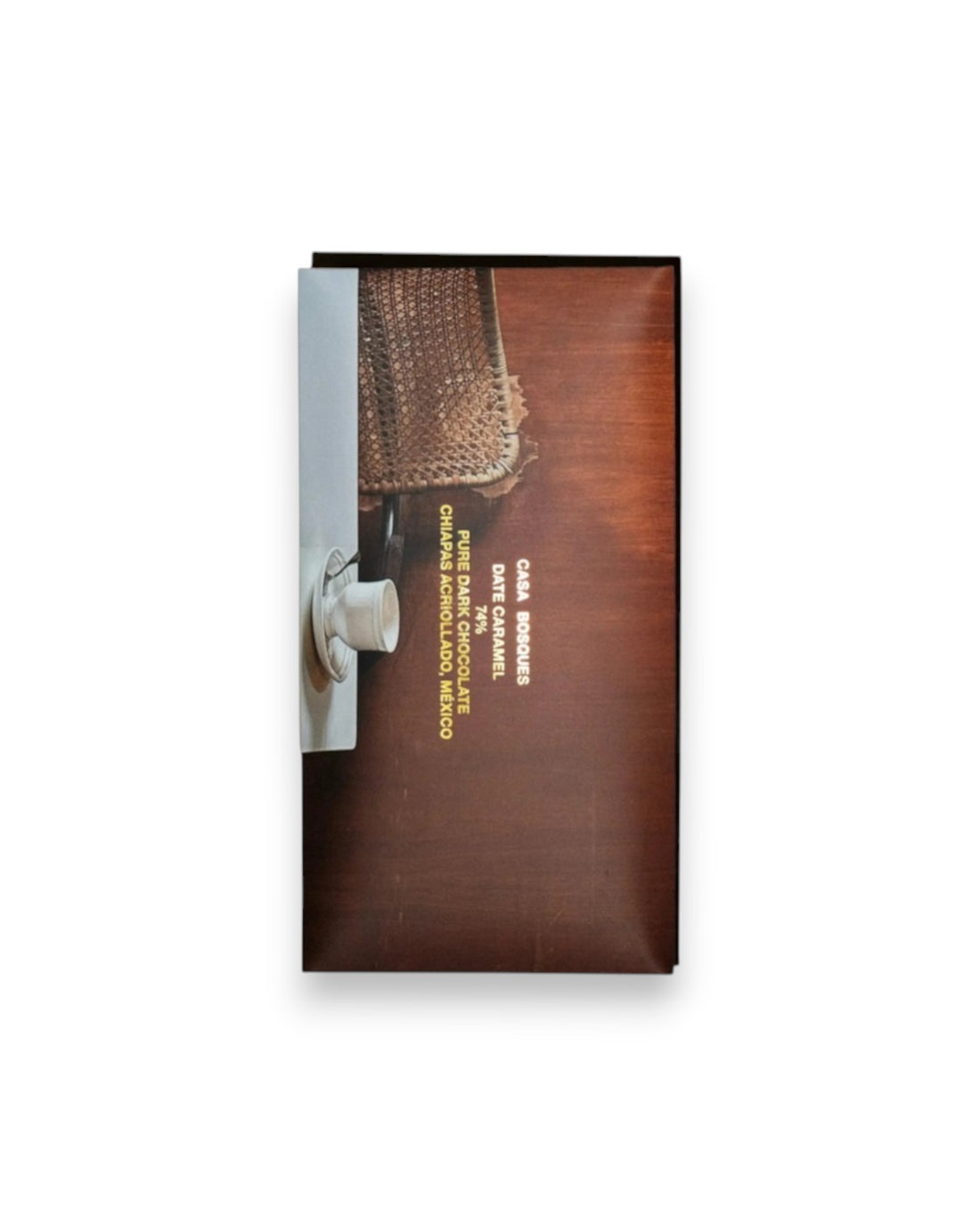A rectangular packaged bar of Casa Bosques chocolate against a solid white background, label reads "CASA BOSQUES DATE CARAMEL 74%" The graphic on packaging is a full bleed photo of a retro style cafe with a woven cane-back chair and a white table with white coffee cup against a brown wooden wall. 