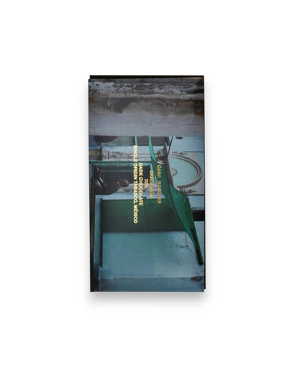 A rectangular packaged bar of Casa Bosques chocolate against a solid white background, label reads "CASA BOSQUES CARDAMOM 70%" The graphic on packaging is a full bleed photo of a colourful building entrance.