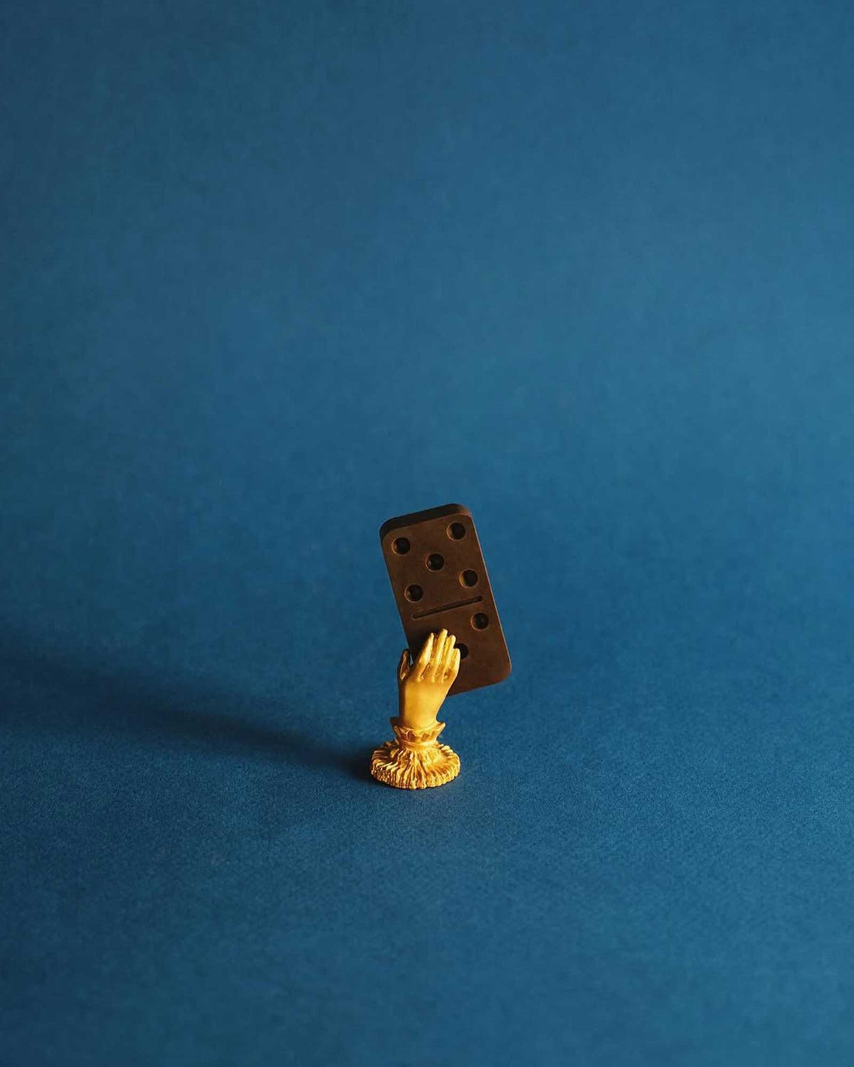 A gold ornamental hand holds a single Casa Bosques chocolate domino piece against a royal blue background. 