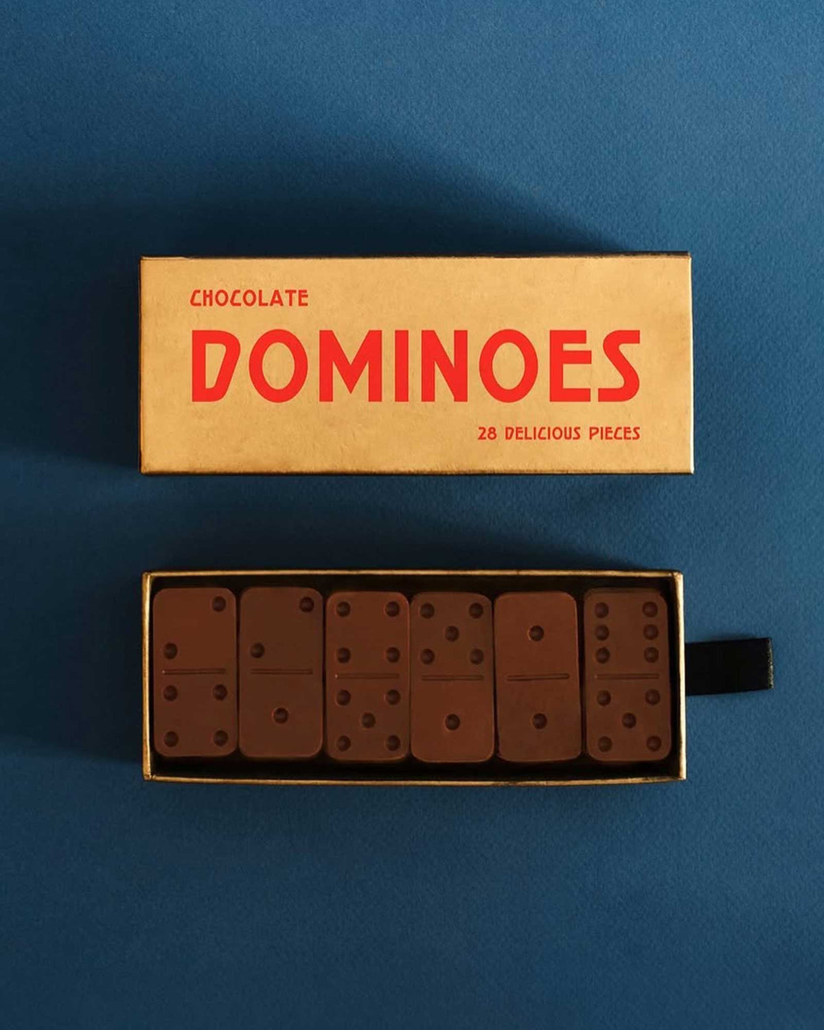 A gold-coloured rectangular boxed set of Casa Bosques chocolate dominoes is displayed with the lid off. The open box shows six chocolate domino pieces. The lid reads "CHOCOLATE DOMINOES 28 DELICIOUS PIECES"