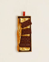 An open box of Casa Bosques chocolate dominoes, four pieces are scattered across the top of the box with a sheet of gold foil organically crumpled underneath the chocolate pieces.  