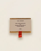 A single rectangular package of Casa Bosques Chocolate Dominoes on a clean off-white background. The package is a light beige/cream color with gold metallic borders and a small red ribbon hanging from the bottom.