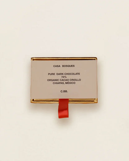 A single rectangular package of Casa Bosques Chocolate Dominoes on a clean off-white background. The package is a light beige/cream color with gold metallic borders and a small red ribbon hanging from the bottom.
