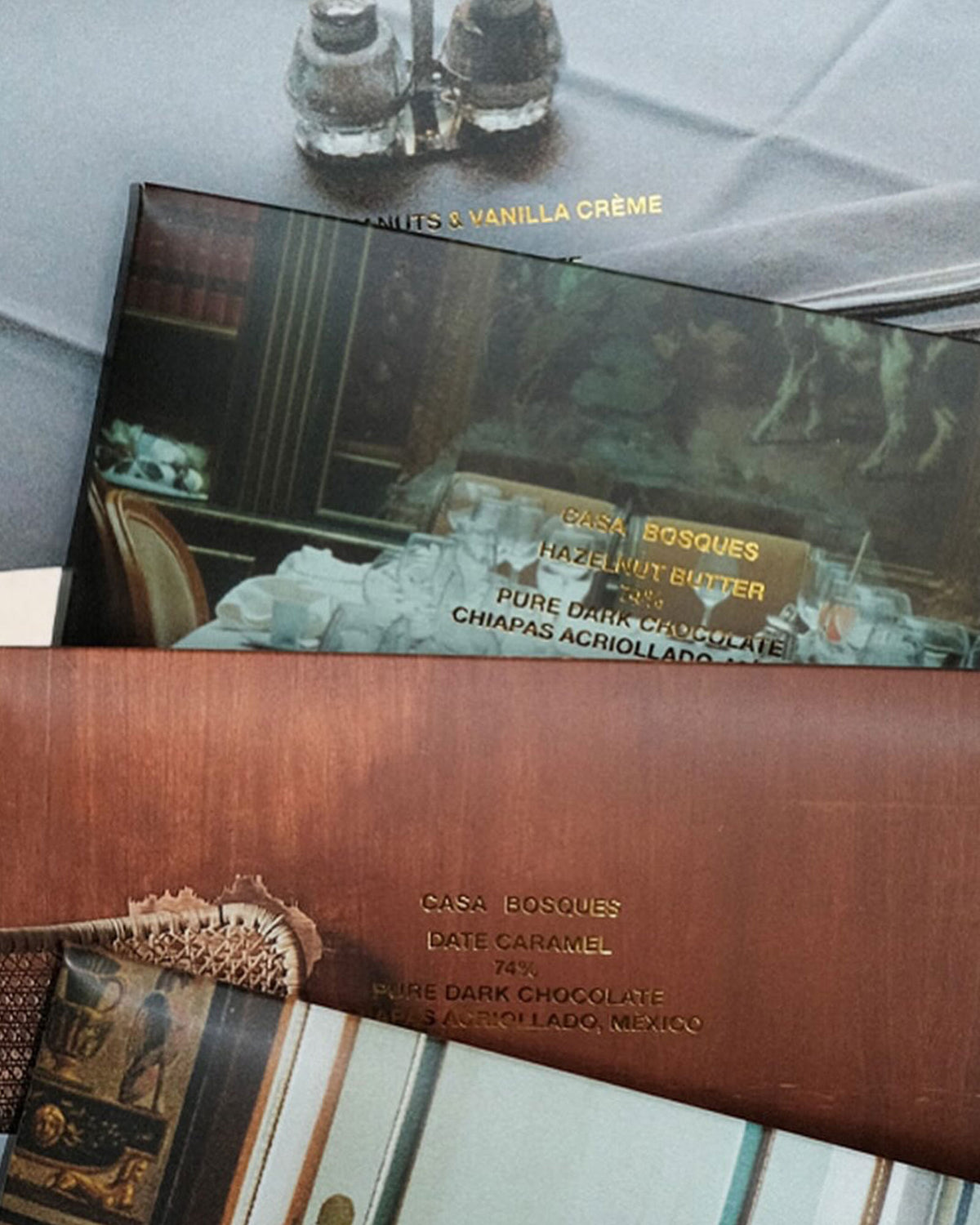 Four rectangular Casa Bosques chocolate bars lay flat on a table, slightly overlapping each other. Each bar is a different flavour with a different photo graphic on outer packaging. The individual flavour and cacao bean location is printed in gold lettering over the photo graphic. 