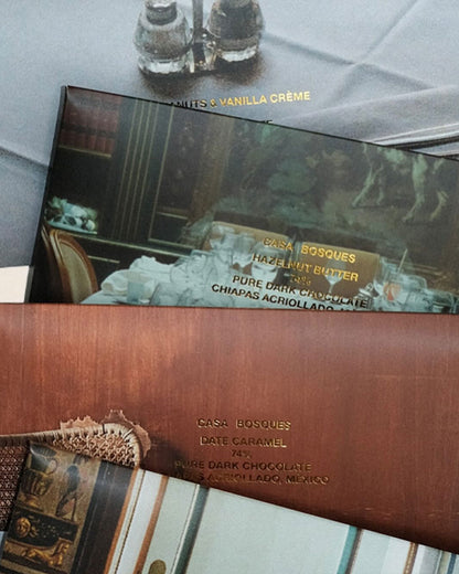 Four rectangular Casa Bosques chocolate bars lay flat on a table, slightly overlapping each other. Each bar is a different flavour with a different photo graphic on outer packaging. The individual flavour and cacao bean location is printed in gold lettering over the photo graphic. 
