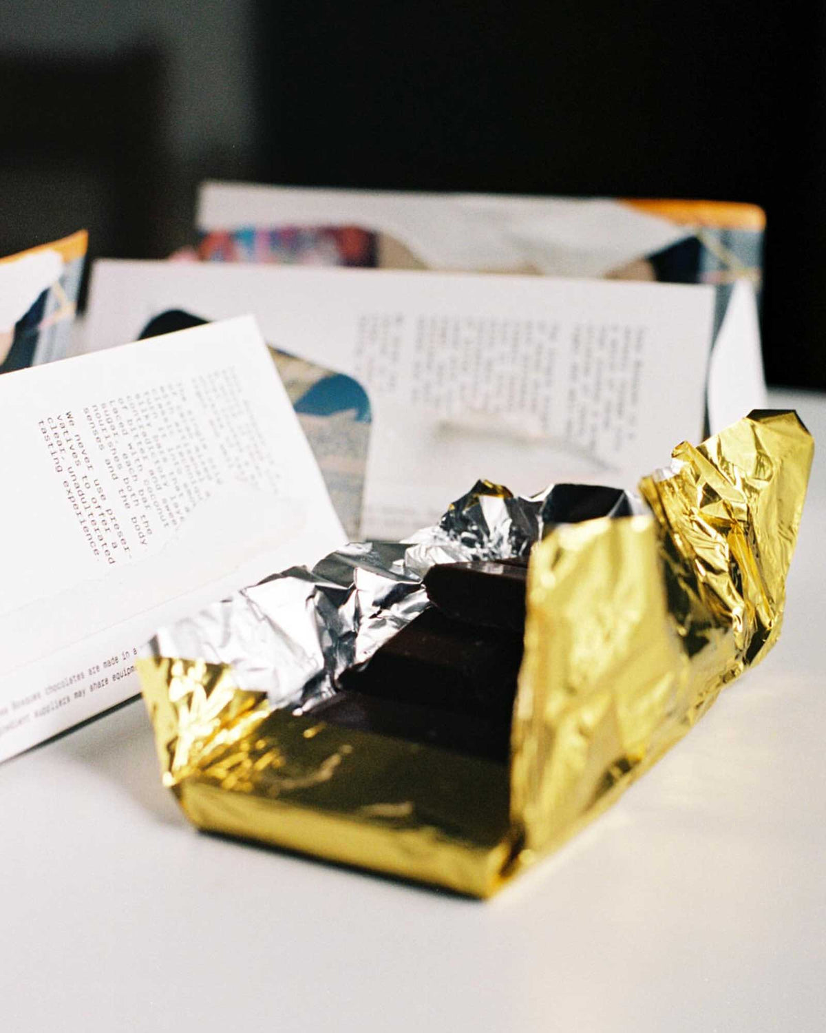 A Casa Bosques rectangular dark chocolate bar is half wrapped in gold and silver foil on a white table with 2 other semi-wrapped bars in background.