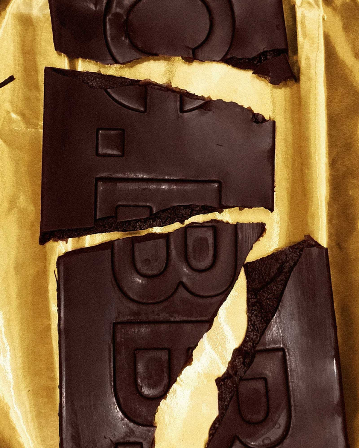 Broken pieces of a rectangular Casa Bosques dark chocolate bar showing a dark cream centre, sitting on a sheet on gold foil. 