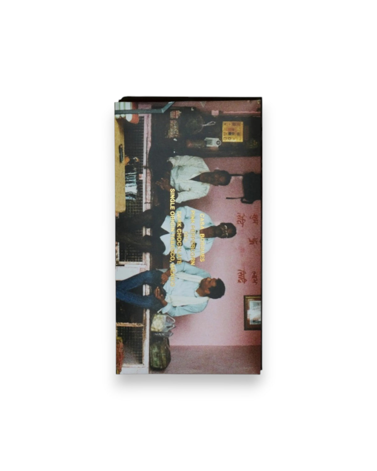 A rectangular packaged bar of Casa Bosques chocolate against a solid white background, label reads "CASA BOSQUES PINK PEPPERCORN 70%" The graphic on packaging is a full bleed photo of 3 Indian men sitting on a bench in front of a pink wall with Indian writing. 