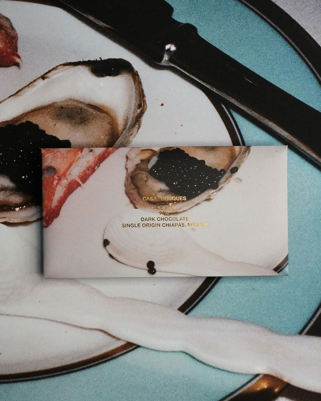 A rectangular packaged bar of Casa Bosques chocolate against a multicoloured background, label reads "CASA BOSQUES SEA SALT 74%" The graphic on packaging is a full bleed photo of a plate with caviar and lobster claw.