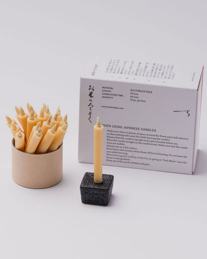 A white product box with Japanese characters and candle burning instructions printed in black ink. The contents of the box are shown next to the box; a cardboard canister with twenty natural-coloured taper candles and a single natural-coloured candle on a square, black stand. 