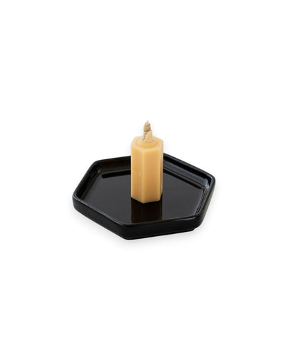 A small honey coloured candle sits on a black hexagon shaped plate on a solid white background. 