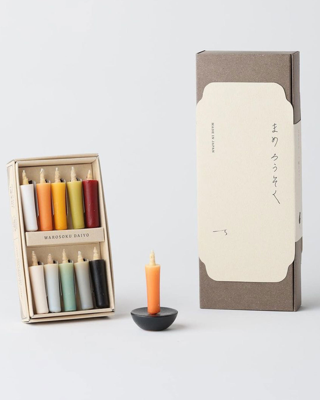 A product shot of a Daiyo mini rice candle set showing a neatly boxed set of 10 earth coloured candles, a small orange candle on a small black circular stand and a tall brown cardboard box with a cream coloured paper slip label. Set on a light grey background. 